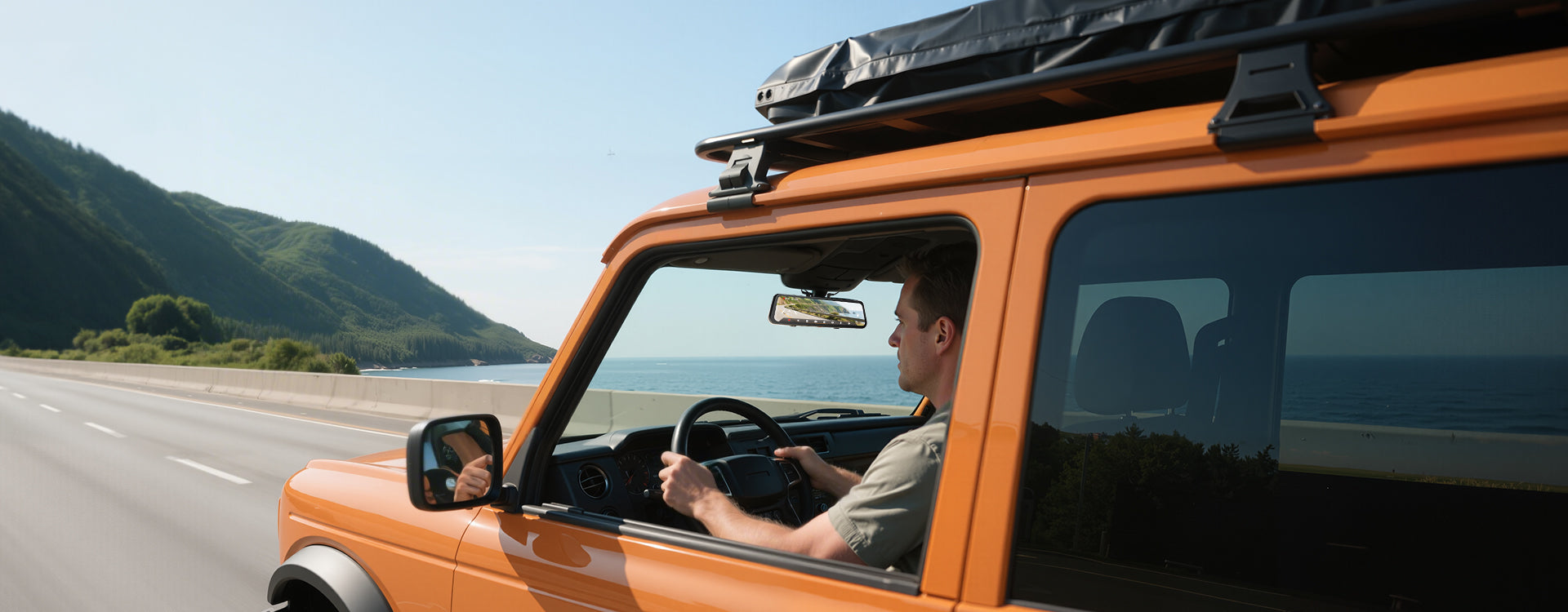 Man driving an orange SUV with a roof rack on a road with mountains in the background
